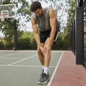 A basketball player holds the front of his knee in discomfort on an outdoor court while experiencing symptoms associated with patellar tendonitis