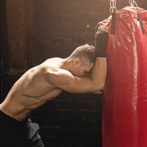 Boxer resting head on punching bag, displaying intense fatigue and signs of overtraining aftermath