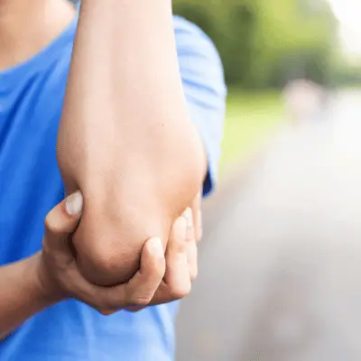 Person grips sore elbow outdoors, showing joint pain focus for sports injury rehab consultation visit