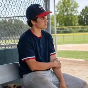 A young baseball player sits in the dugout holding his arm near the elbow while resting because of possible little league elbow discomfort