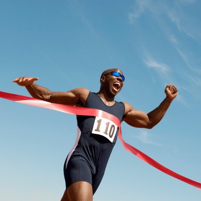 Sprinter wearing race bib bursts through red finish ribbon, raising arms in victory under sky