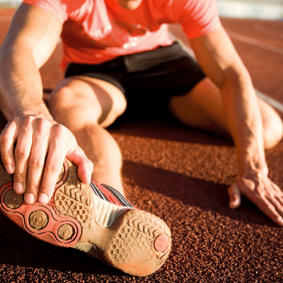 Runner sits on track, stretching hamstring and calf by reaching forward to hold shoe sole