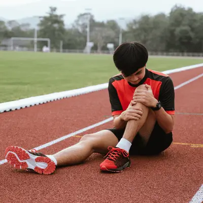 Teen runner sits on track, holding knee, considering runners knee rehab after overuse pain today